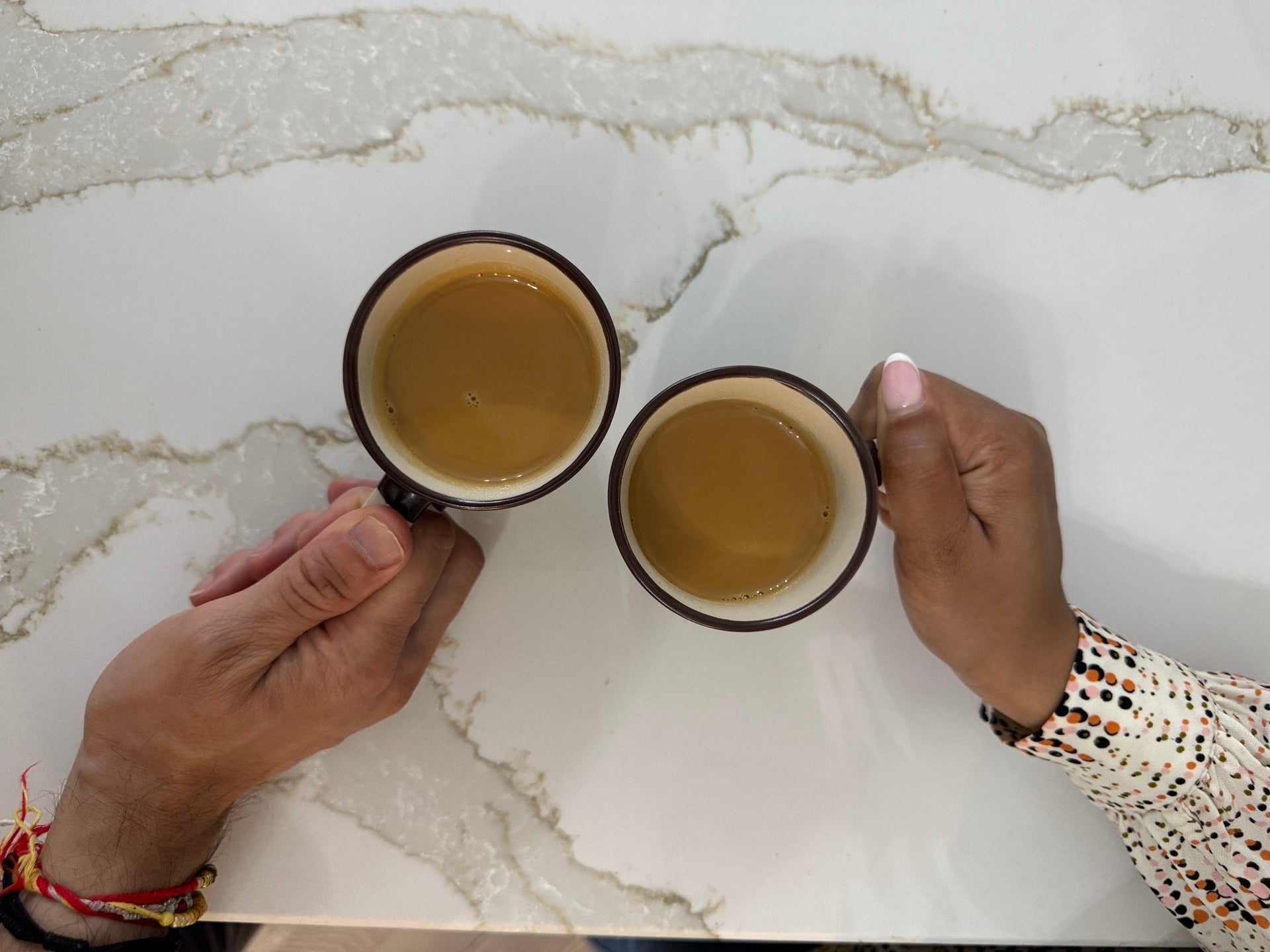 Two hands holding cups of chai on a marble surface